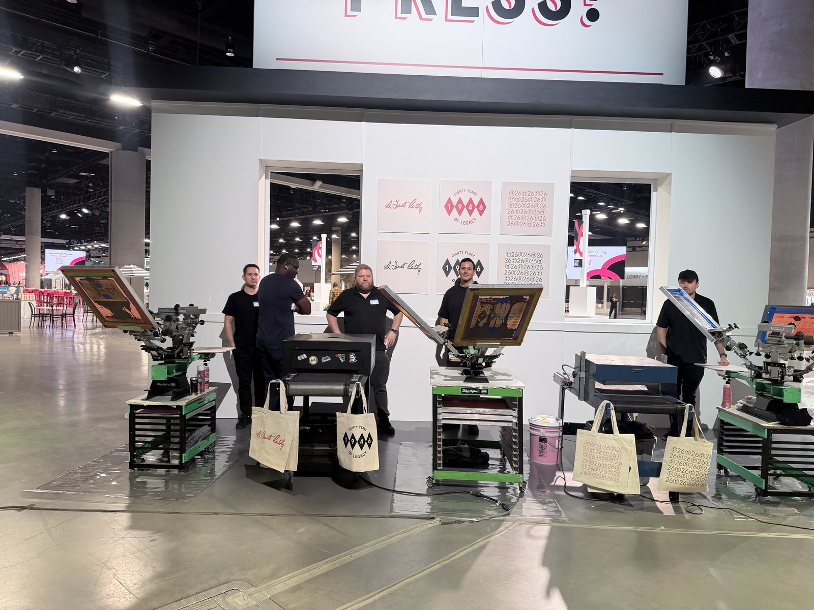 Wide convention floor booth with multiple staffed stations and script-lettered tote bags displayed on the equipment