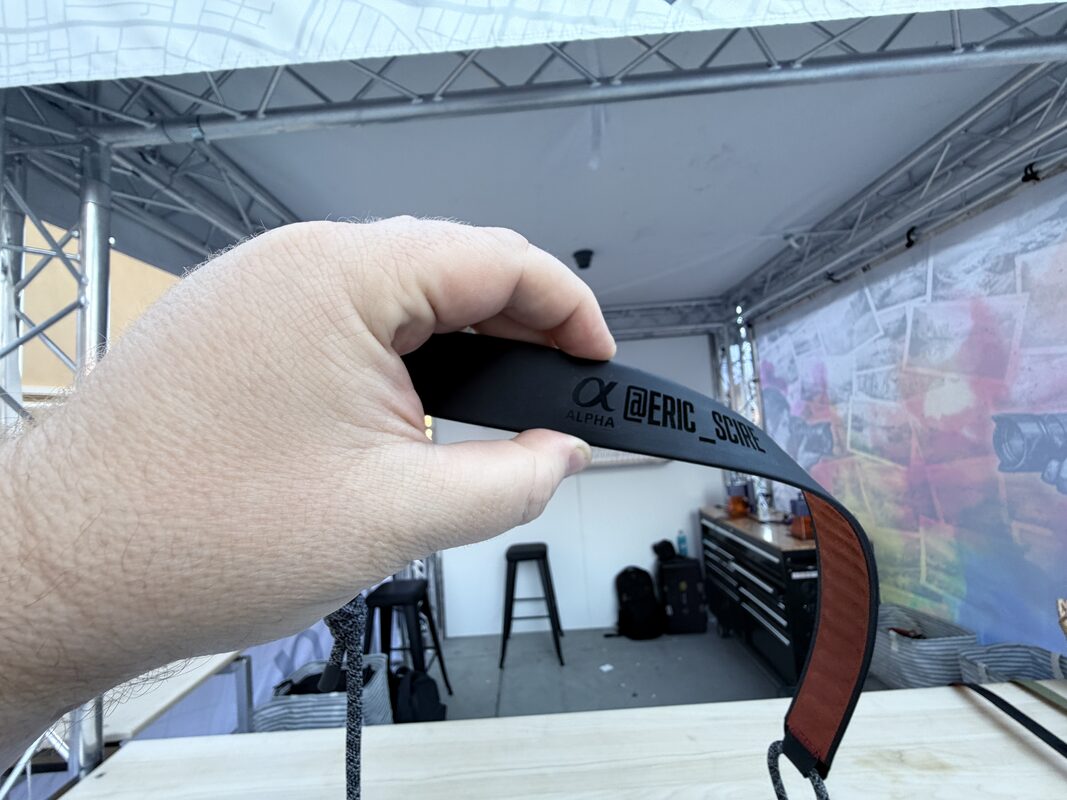 Hand holding a black camera strap personalized with a guest name in bold lettering at an outdoor festival booth